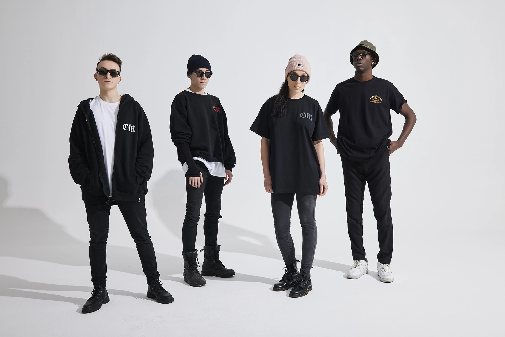 Four fashion models dressed in black contemporary outfits posing together in a minimalist studio setting.