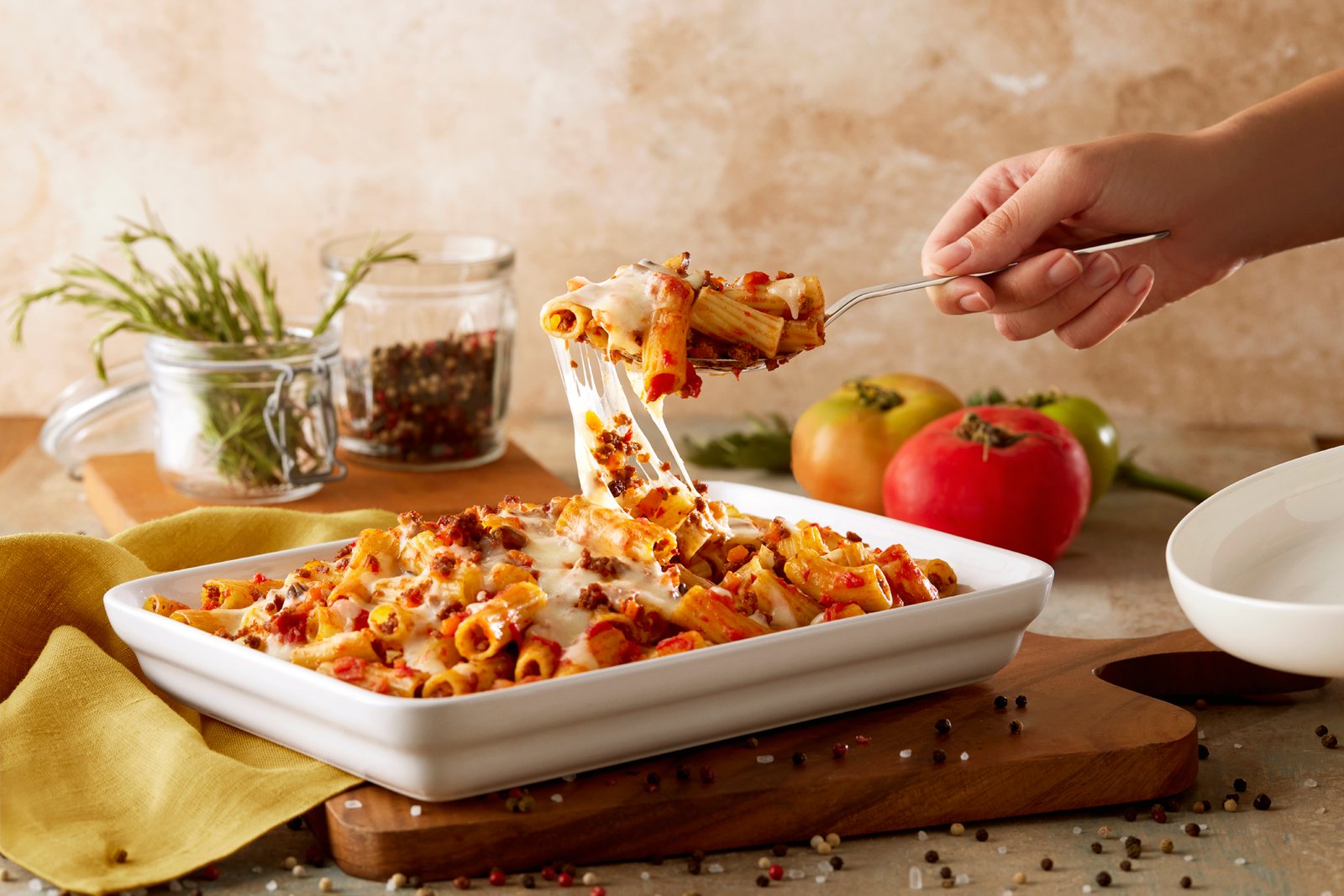 Professional commercial food advertising photograph by Sinan Niyazi Kutsal capturing a hand using a spoon to lift a cheesy portion of baked Pastavilla Rigatoni pasta with an impressive cheese pull effect over a ceramic dish.