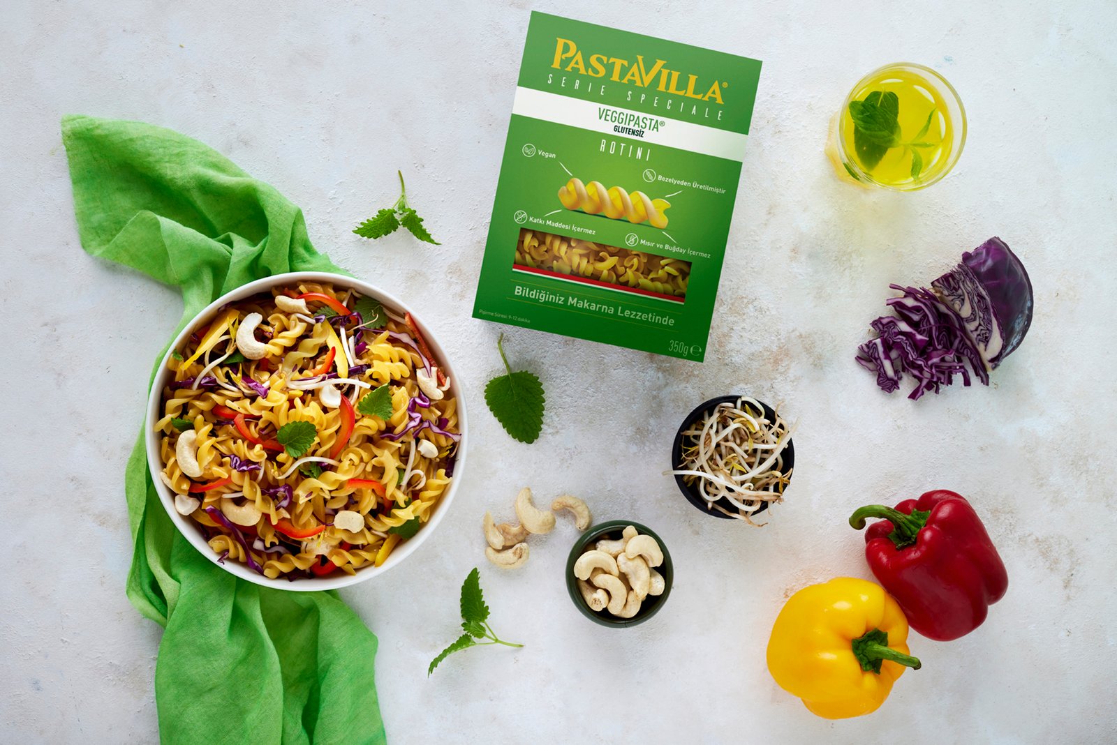 Professional commercial food advertising photograph by Sinan Niyazi Kutsal capturing a flat lay of a Pastavilla Veggipasta box next to a colorful bowl of gluten-free rotini salad with cashews and fresh vegetables on a white stone background.