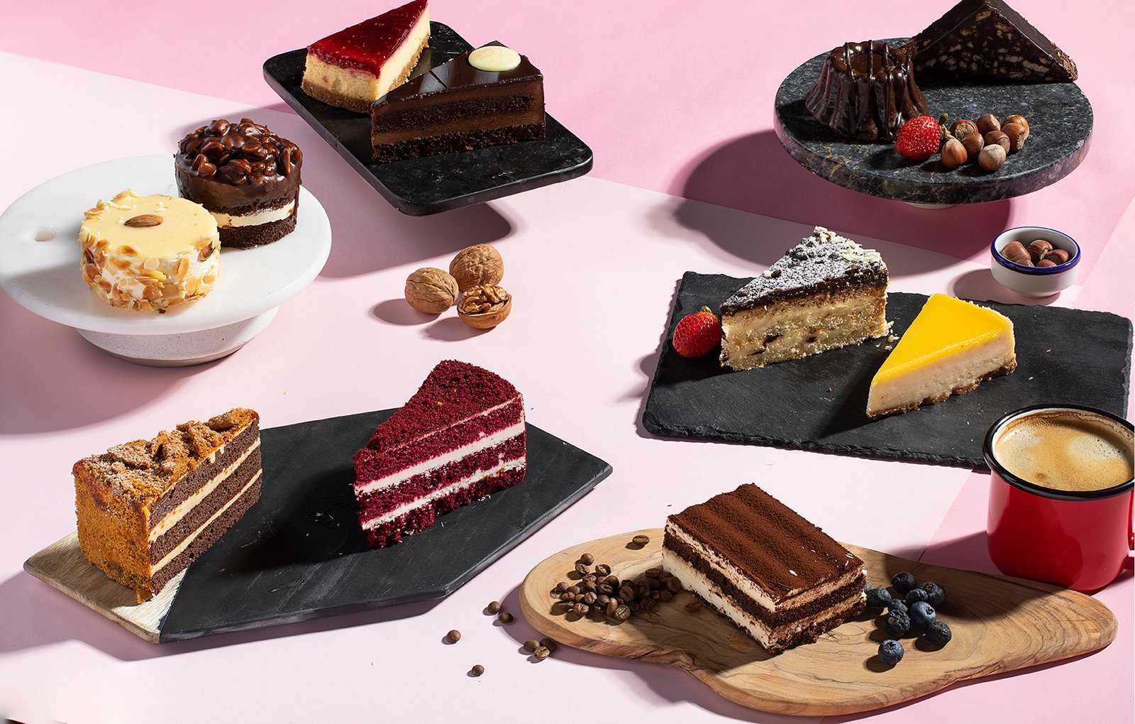 Professional commercial food advertising photograph by Sinan Niyazi Kutsal capturing a colorful flat lay display of various gourmet cake slices, fresh berries, walnuts, and a red cup of coffee on a pink and white surface.