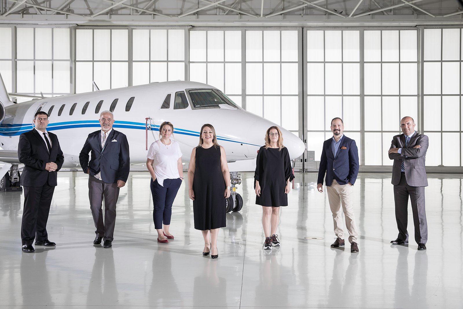 Professional commercial corporate photograph by Sinan Niyazi Kutsal, capturing a diverse group of corporate executives in formal attire posing in front of a white private jet airplane inside a large hangar.