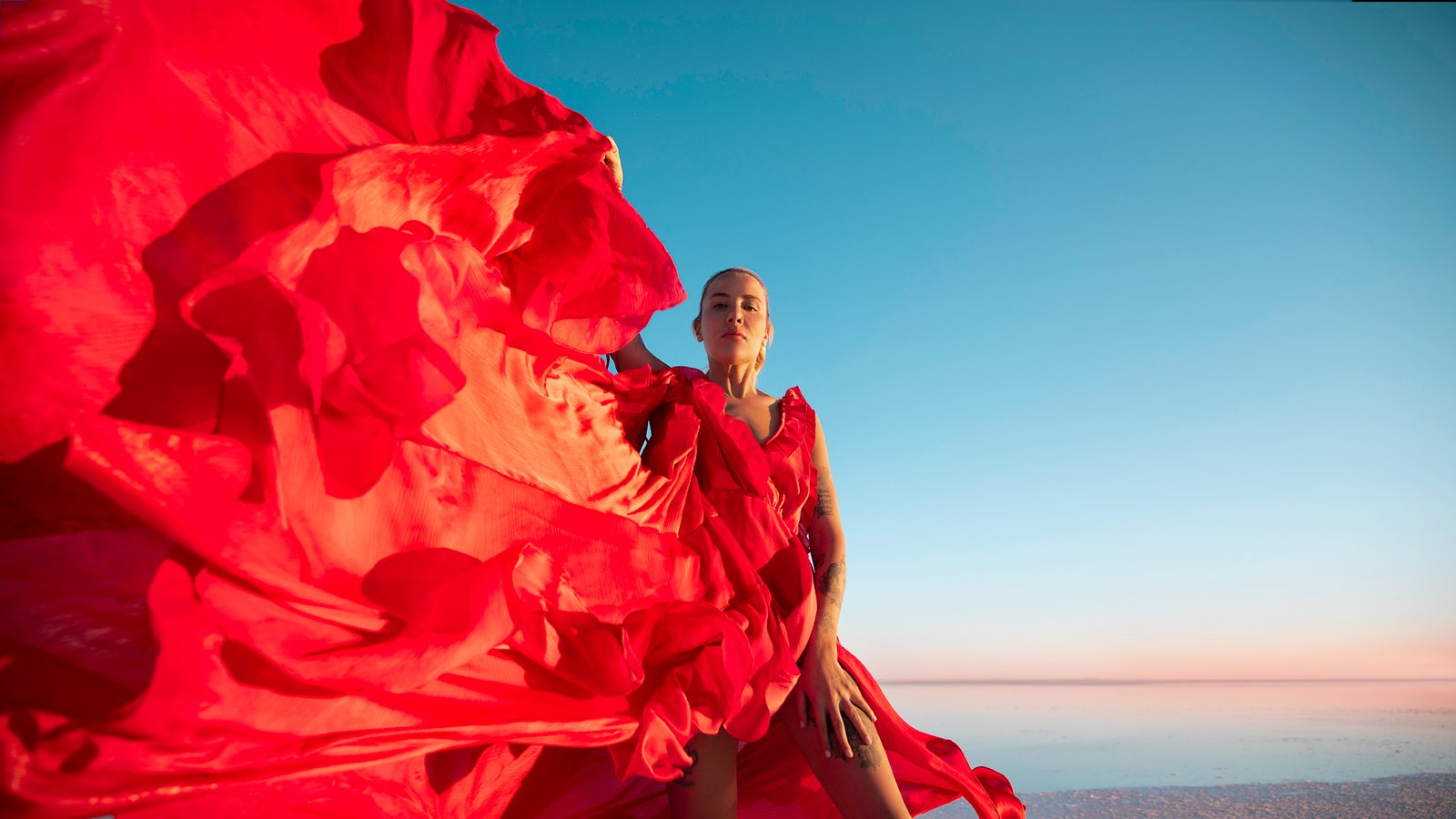 Professional cinematic fine art fashion photographs by Sinan Niyazi Kutsal capturing a female model in a magnificent flowing red dress that blows across the vast, mirroring salt flats of Tuz Golu in Aksaray, Turkey under a clear blue pastel sky.