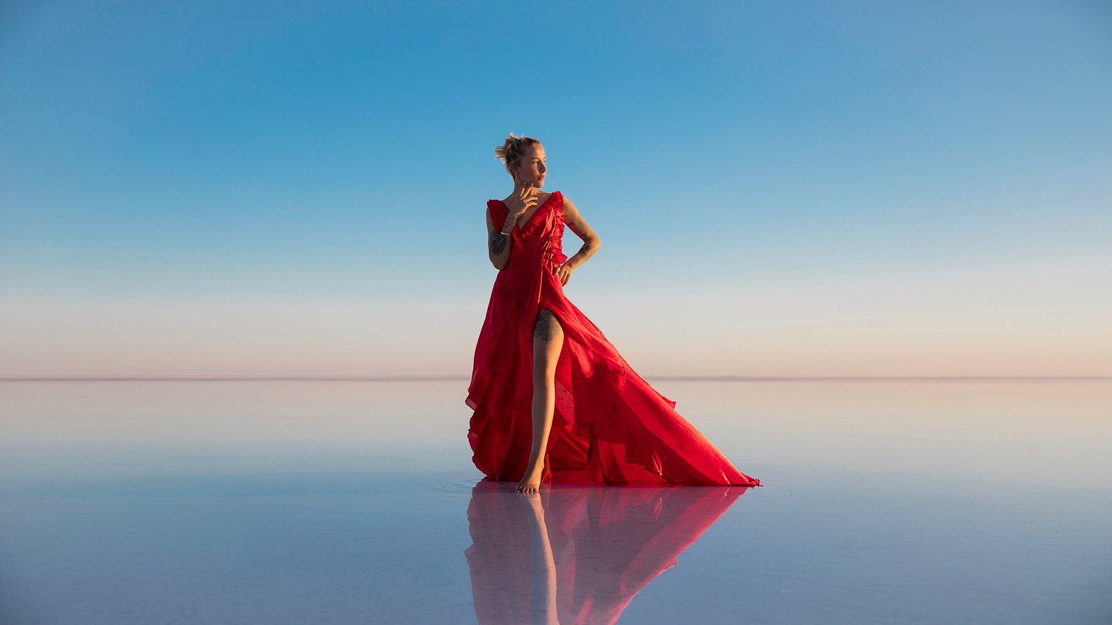 Professional cinematic fine art fashion photographs by Sinan Niyazi Kutsal capturing a female model in a magnificent flowing red dress that blows across the vast, mirroring salt flats of Tuz Golu in Aksaray, Turkey under a clear blue pastel sky.