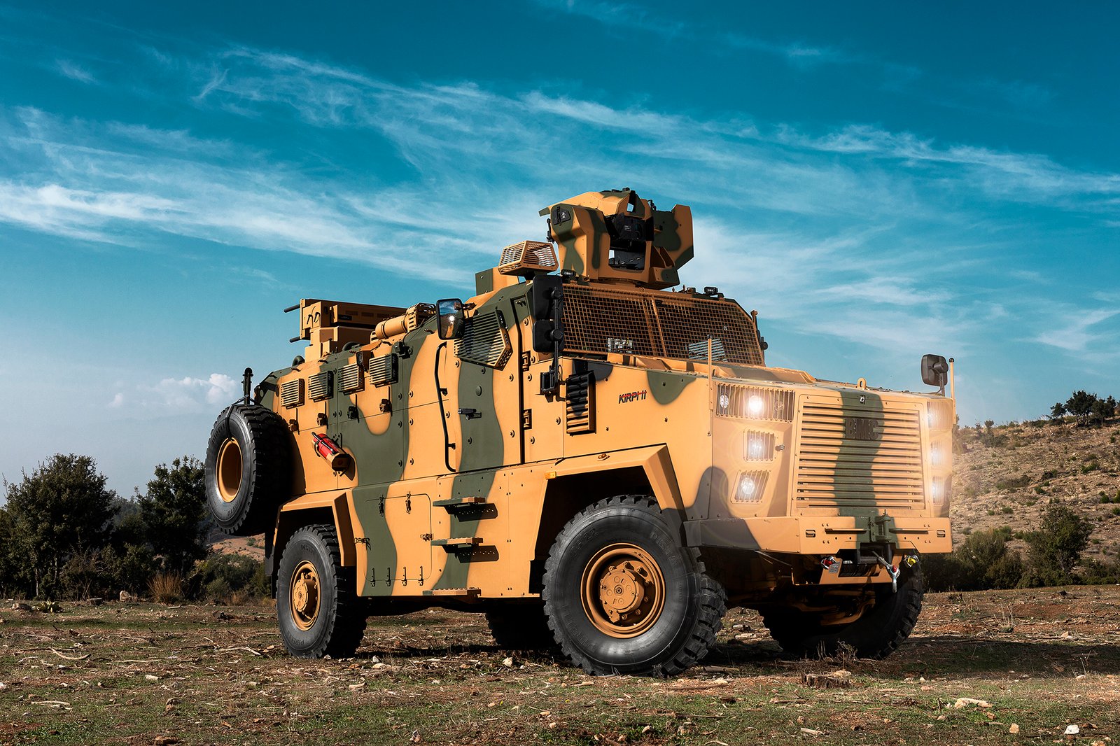 Professional commercial defense industry photograph by Sinan Niyazi Kutsal capturing a camouflage BMC Kirpi II heavy armored MRAP vehicle in a rugged dirt field in Izmir under a bright blue sky.
