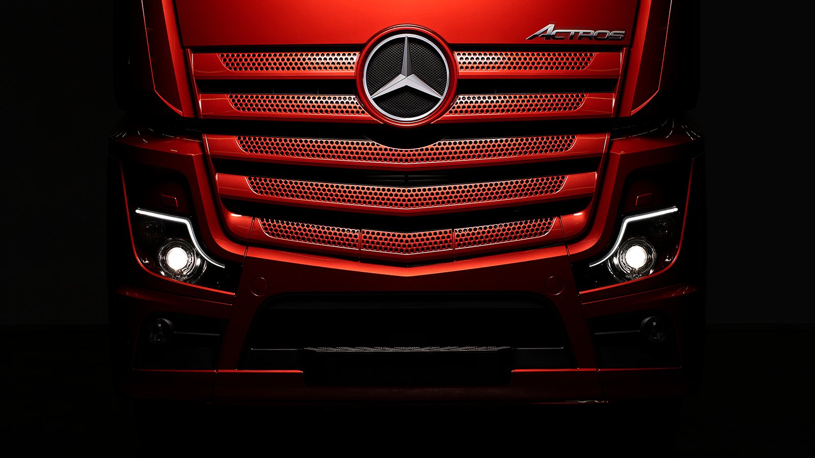A dramatic commercial close-up photograph by Sinan Niyazi Kutsal capturing the center front of a metallic red Mercedes-Benz Actros truck, showcasing the gleaming chrome star emblem and illuminated headlights against a deep black background.