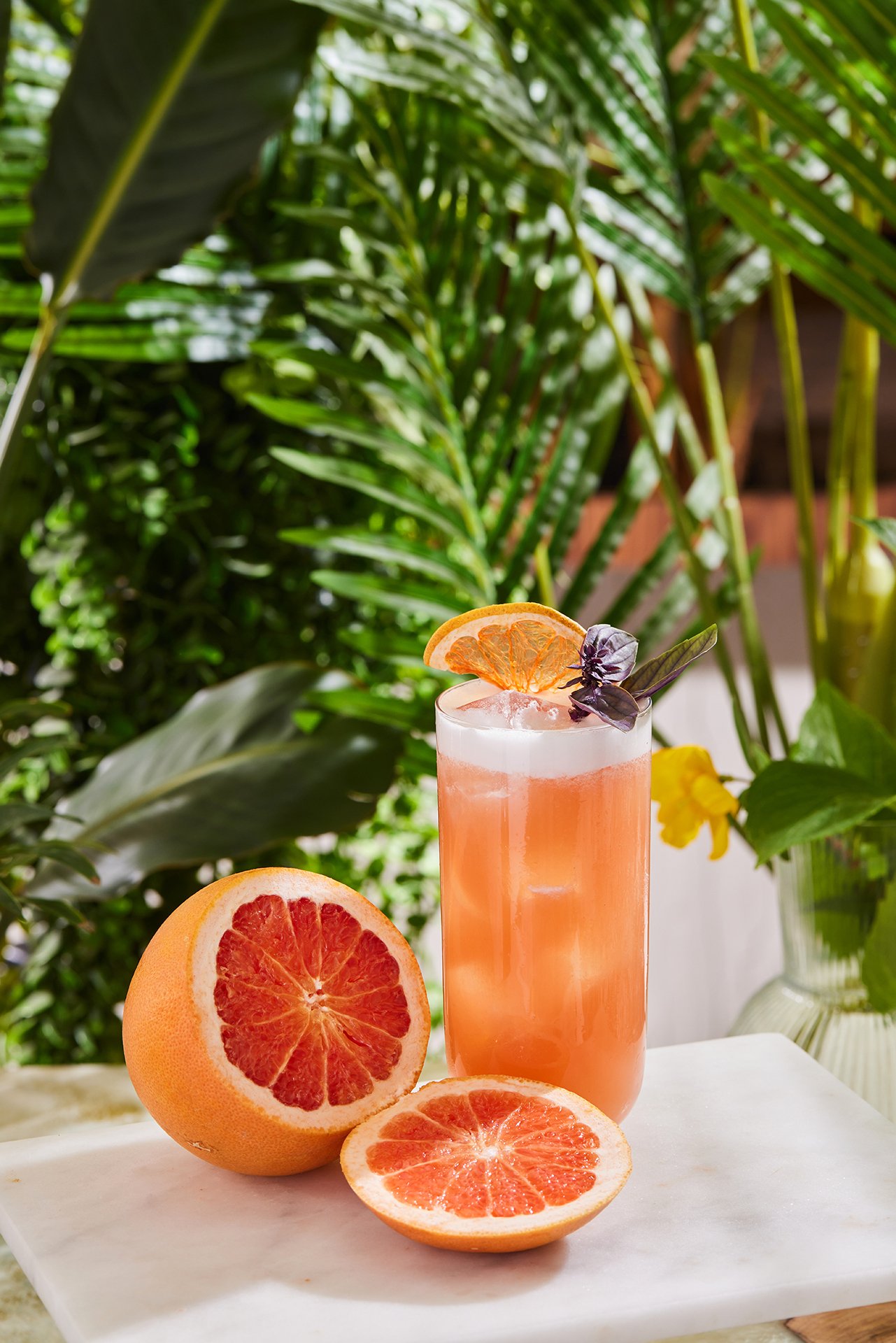 A vibrant and fresh commercial beverage photograph by Sinan Niyazi Kutsal for Barley Brasserie, featuring a tall pink cocktail garnished with a dried orange wheel next to sliced fresh ruby red grapefruits on a white marble board.