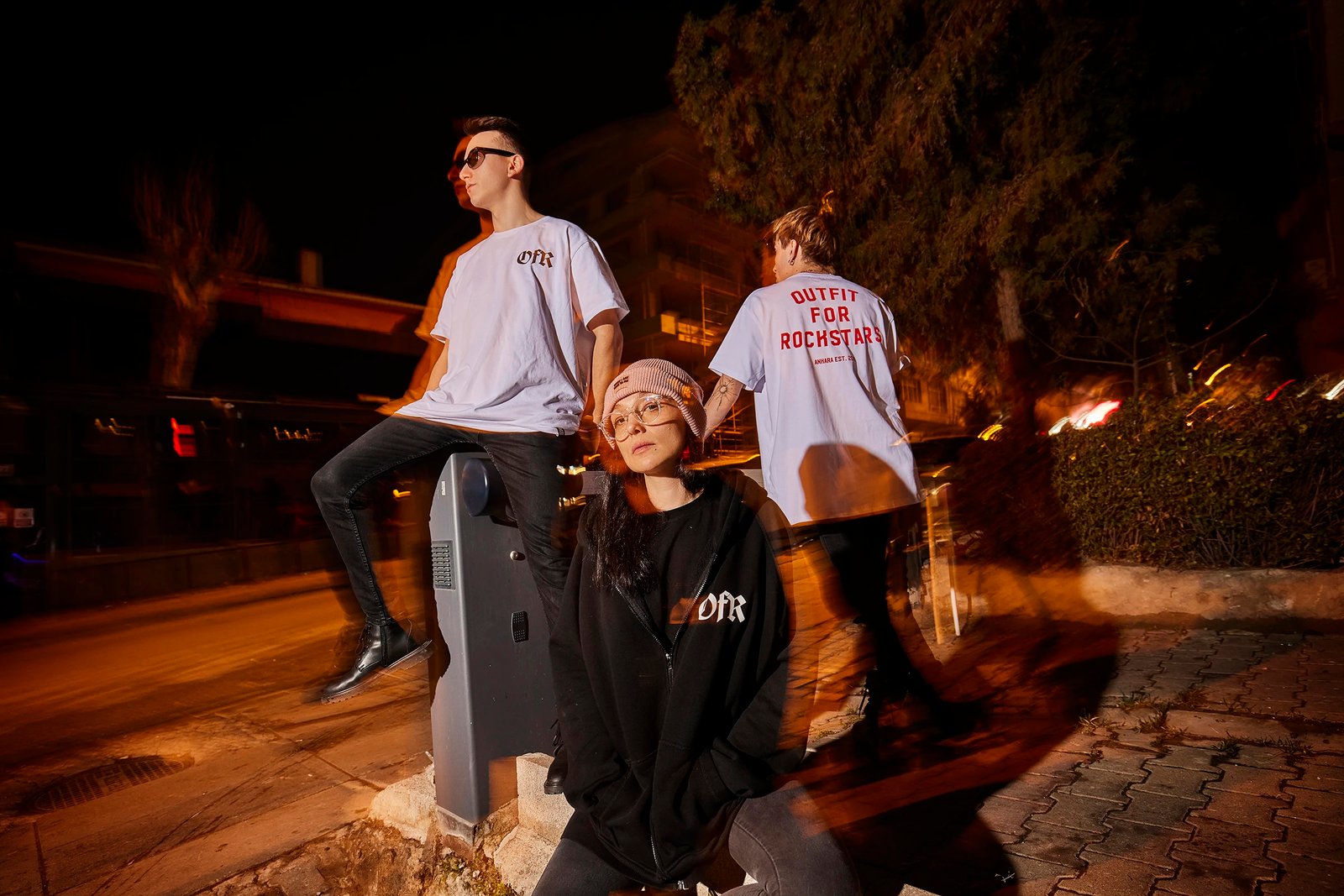 A night streetwear fashion photoshoot by Sinan Niyazi Kutsal featuring three models posing on a city street. One model wears a black oversized OFR zipped hoodie and pink beanie, while two models in the background wear white graphic t-shirts displaying 'OFR' and 'Outfit For Rockstars' in red gothic font, captured with a dynamic long exposure motion blur effect.