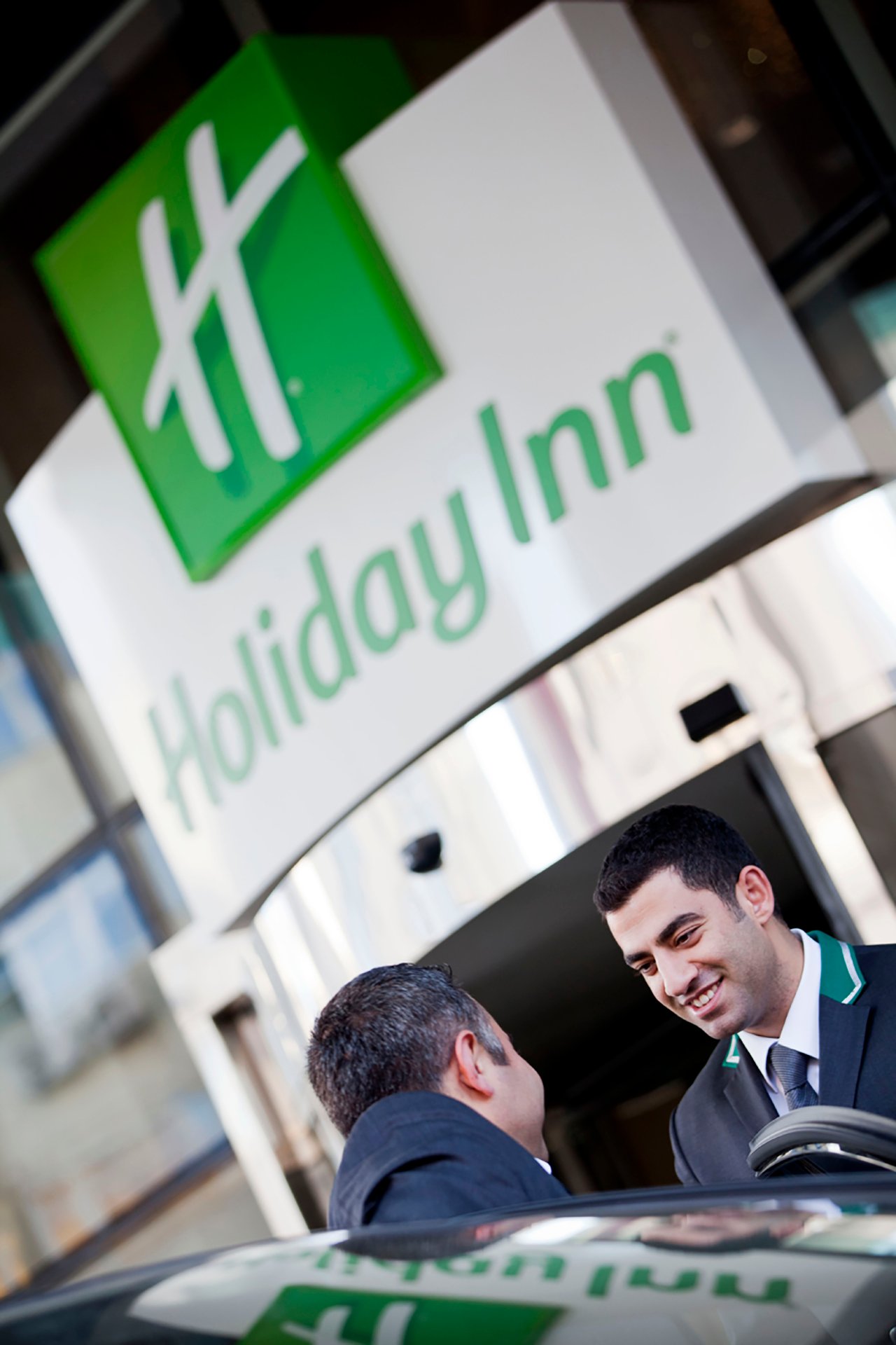 A professional commercial hospitality photograph by Sinan Niyazi Kutsal showcasing a smiling Holiday Inn hotel employee greeting a guest beneath the corporate green and white Holiday Inn sign.