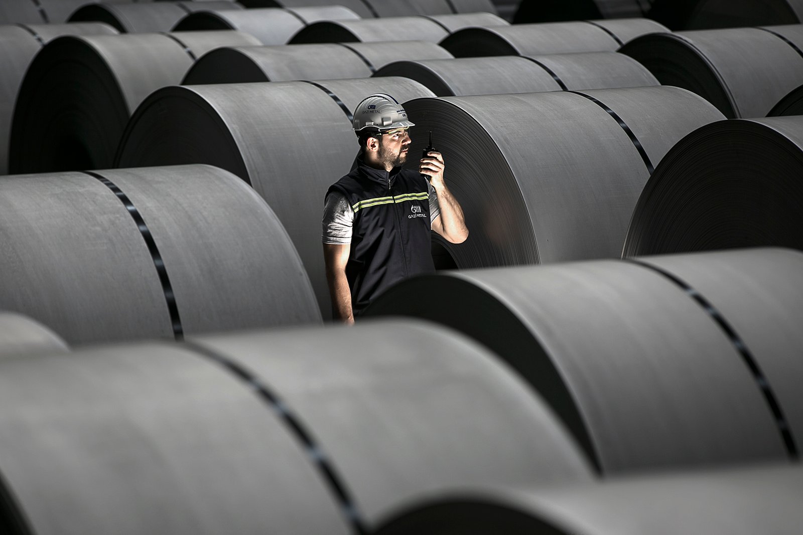 Professional commercial industrial photographs by Sinan Niyazi Kutsal for Gazi Metal, capturing manufacturing plant operators inspecting immense rolls of steel sheeting using radio communication inside a Sakarya warehouse.