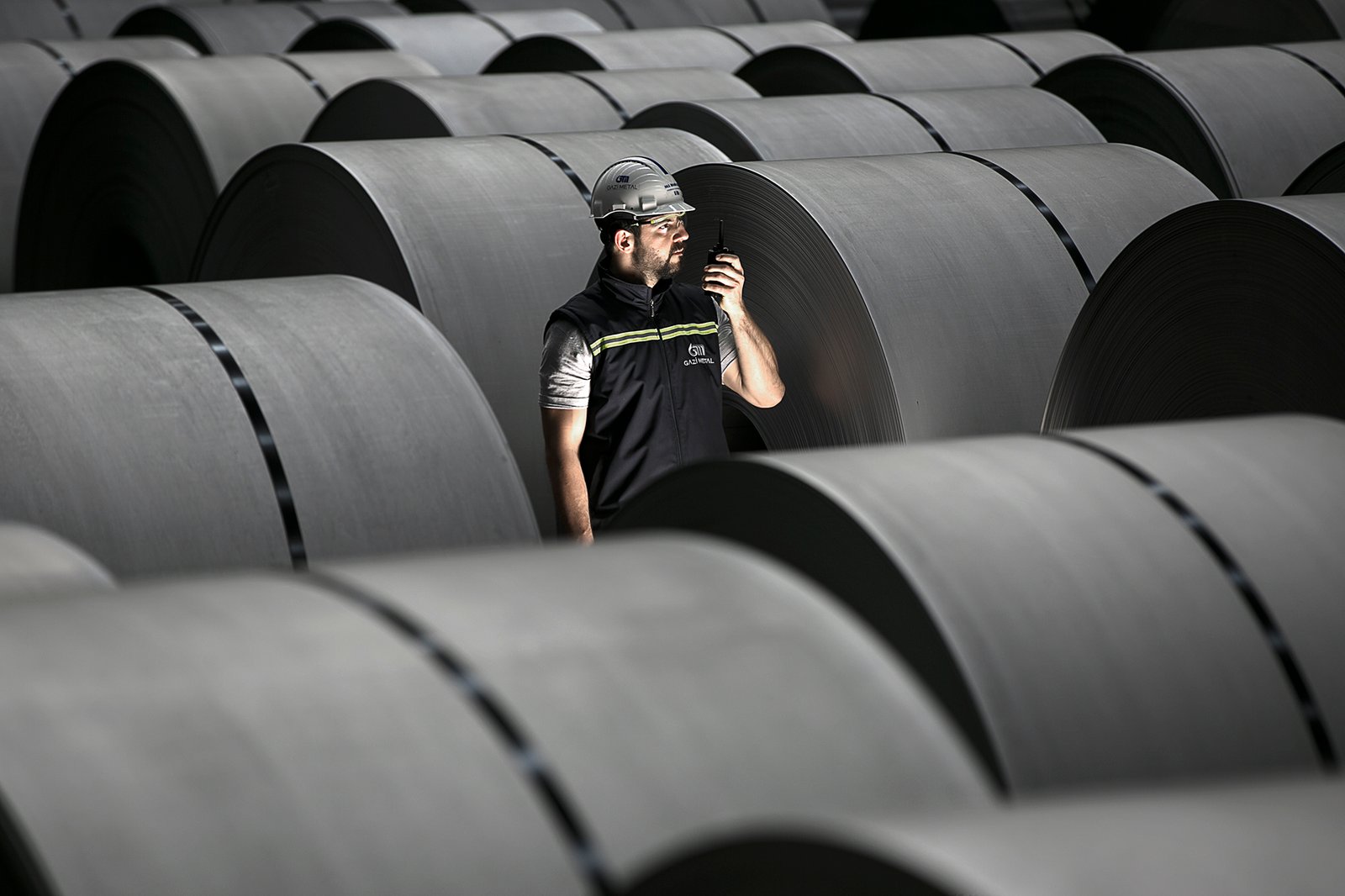 Professional commercial industrial photographs by Sinan Niyazi Kutsal for Gazi Metal, capturing a factory supervisor with a walkie-talkie among massive rows of steel coils, a worker framed inside a steel coil cylinder under blue light, and a silhouette of three workers under flags.