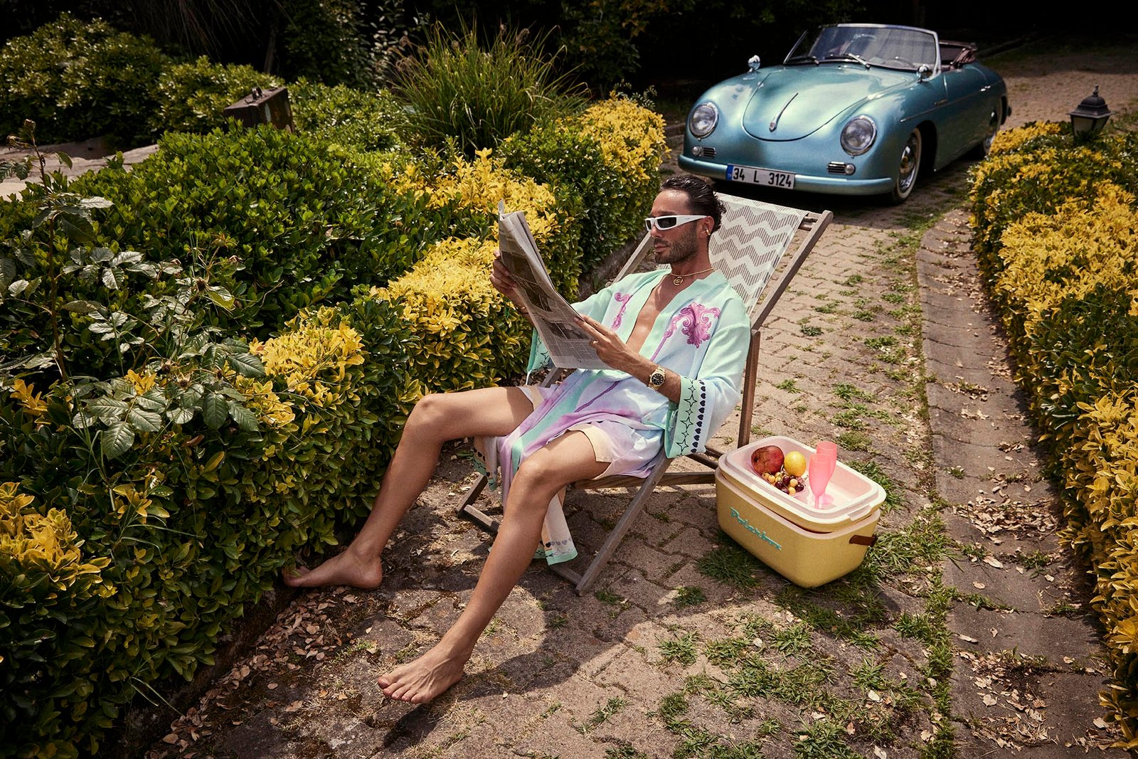 A luxury resortwear fashion photo by Sinan Niyazi Kutsal featuring male model Batuhan Baskan wearing a mint green and purple printed silk robe and white sunglasses, relaxing on a deck chair in a lush green Zekeriyakoy garden with a vintage metallic cyan Porsche 356 Speedster parked in the background.