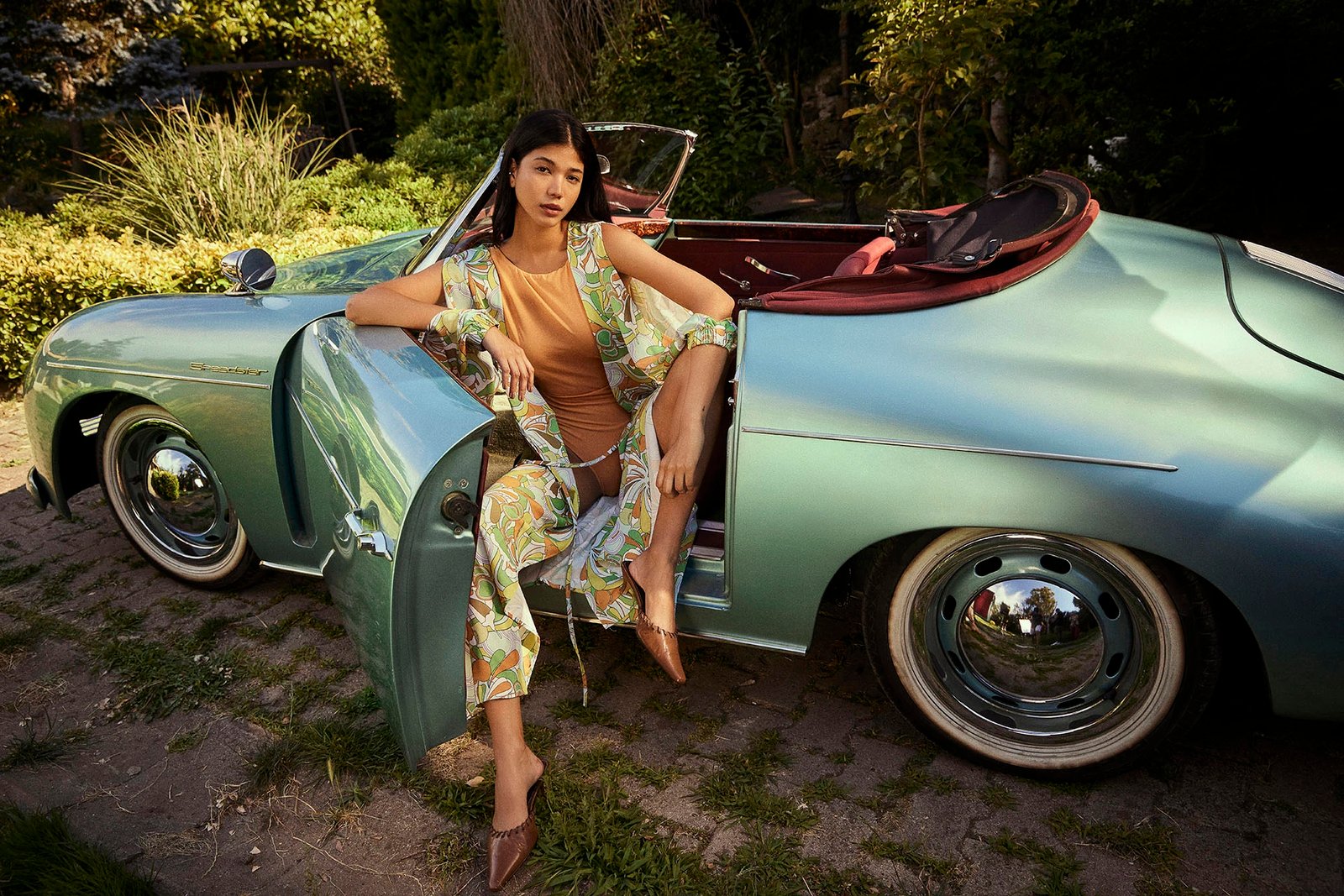 A commercial fashion photograph by Sinan Niyazi Kutsal showing an attractive female model in a green and orange retro printed silk robe sitting on the open door of a vintage metallic cyan Porsche convertible car.