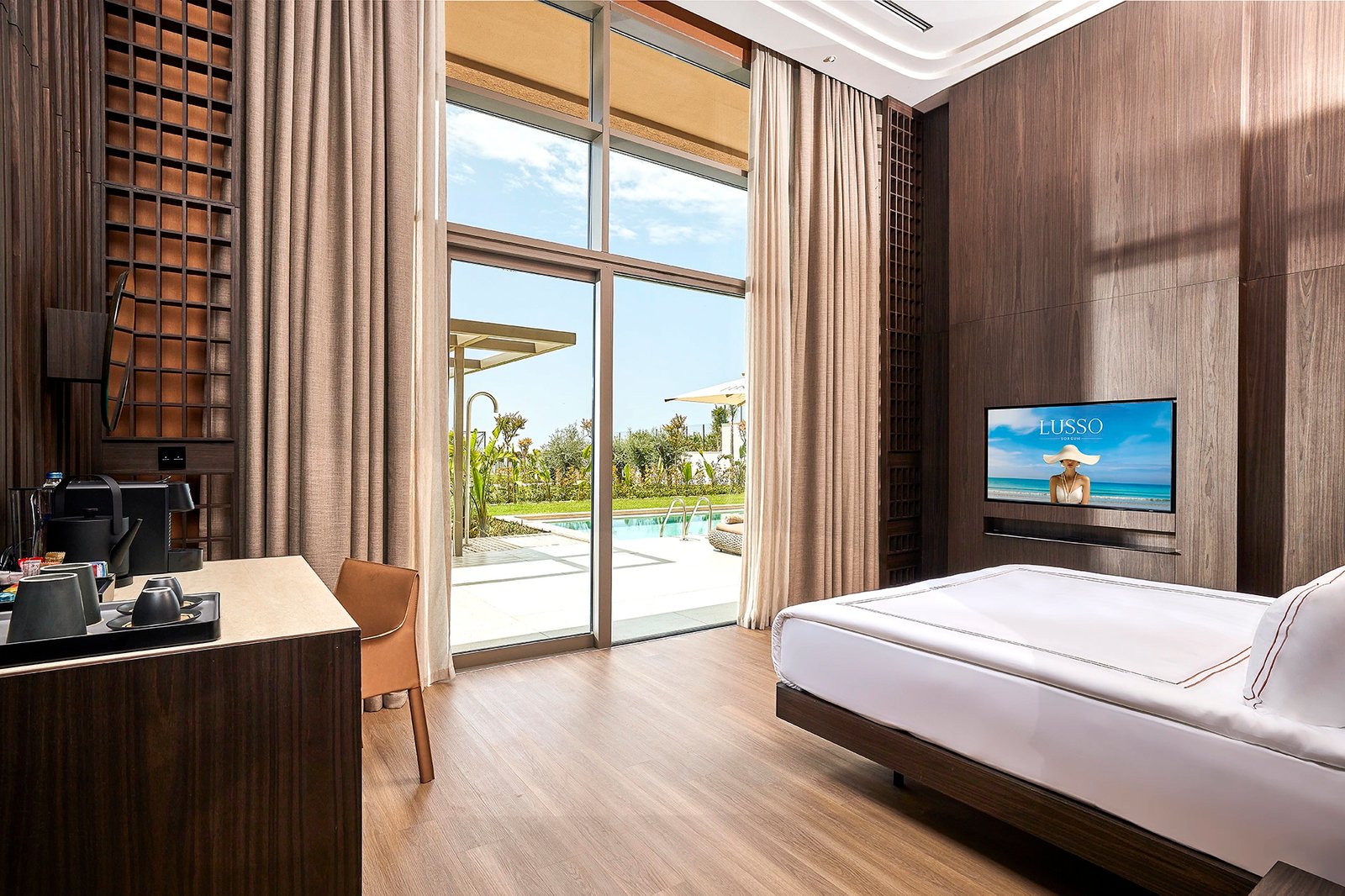 A commercial interior photograph by Sinan Niyazi Kutsal for Lusso Sorgun, highlighting a high-ceiling hotel bedroom with vertical wood screens, a white bed, and grand sliding doors revealing an outdoor private pool area.