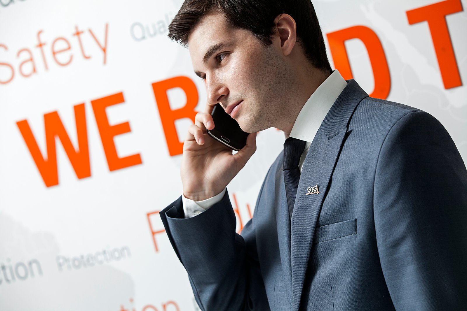 Professional commercial corporate photograph by Sinan Niyazi Kutsal for SGS, capturing a businessman in a blue suit with an SGS pin talking on a mobile phone in front of a white wall with corporate values text.