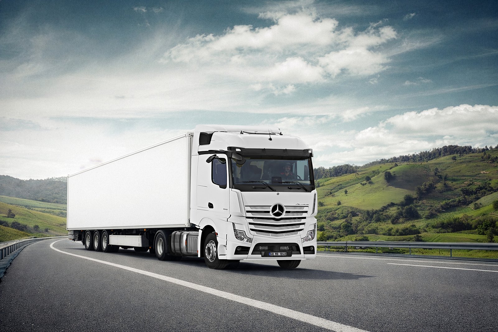 A commercial high-angle automotive photograph by Sinan Niyazi Kutsal capturing a large white Mercedes-Benz Actros semi-truck and box trailer driving along a curved mountain highway layout.