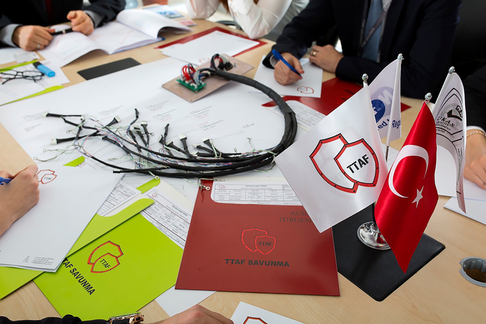 Professional commercial corporate photographs by Sinan Niyazi Kutsal for TTAF Savunma, capturing a soldier with a rifle and tech helmet next to laboratory engineers, and a tabletop setup of corporate folders, cable harnesses, and Turkish desk flags.