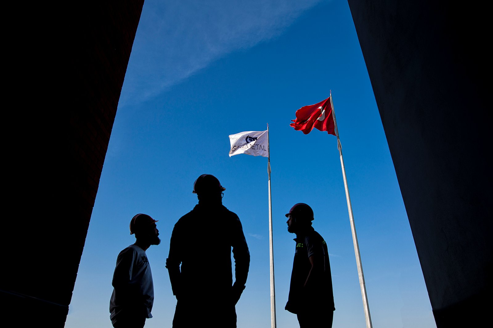 Professional commercial industrial photographs by Sinan Niyazi Kutsal for Gazi Metal, capturing a factory supervisor with a walkie-talkie among massive rows of steel coils, a worker framed inside a steel coil cylinder under blue light, and a silhouette of three workers under flags.