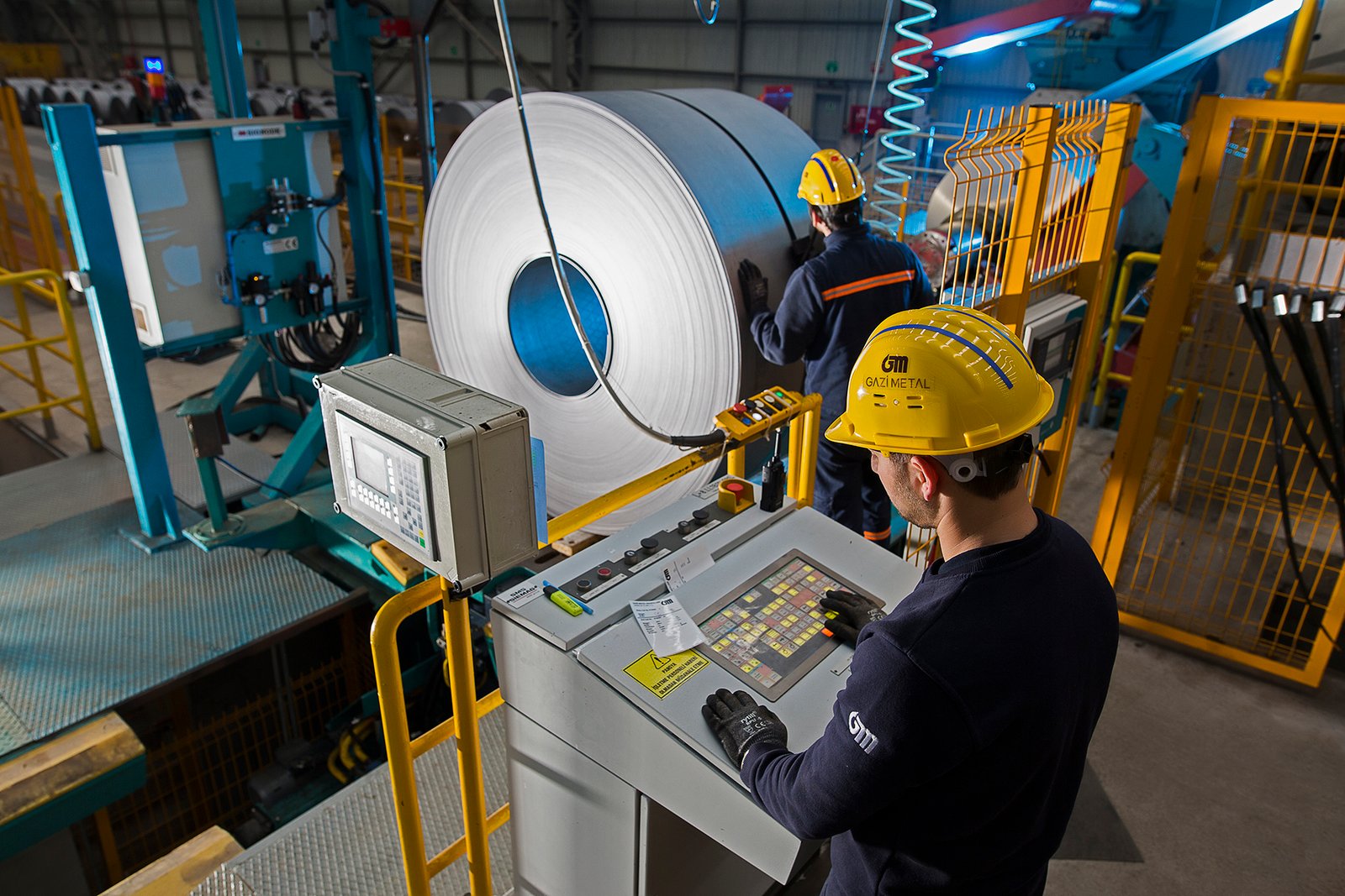 Professional commercial industrial photographs by Sinan Niyazi Kutsal for Gazi Metal capturing plant engineers operating automated console panels, a large overhead crane hoist picking up a steel coil, and rows of metal sheets in a warehouse.
