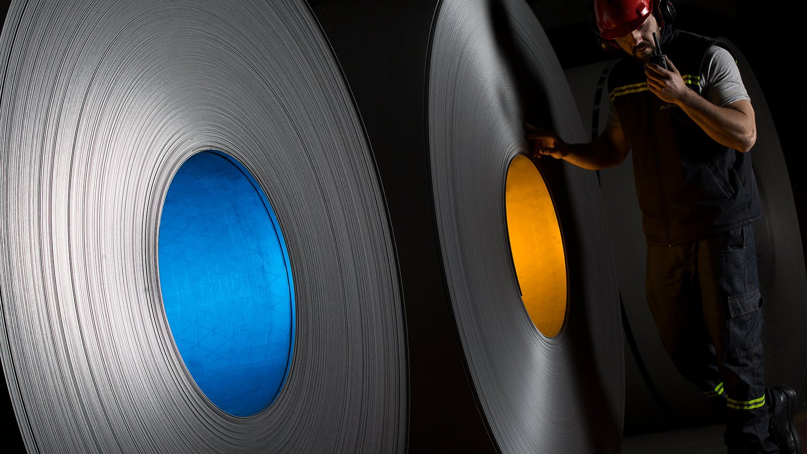 Professional commercial industrial photographs by Sinan Niyazi Kutsal for Gazi Metal, capturing manufacturing plant operators inspecting immense rolls of steel sheeting using radio communication inside a Sakarya warehouse.