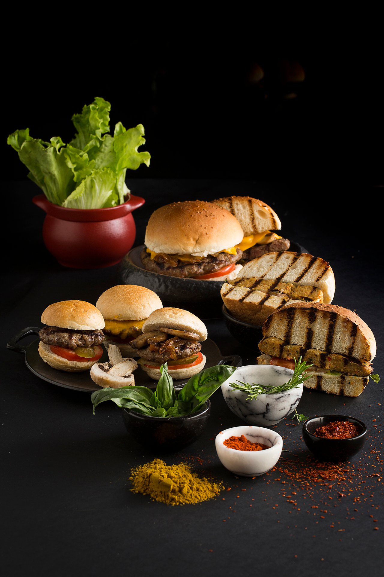 Professional commercial food advertising photographs by Sinan Niyazi Kutsal showcasing a rich spread of gourmet mini slider burgers with spices, and a close-up of a charcoal black bun cheeseburger with seasoned french fries on a wooden board.