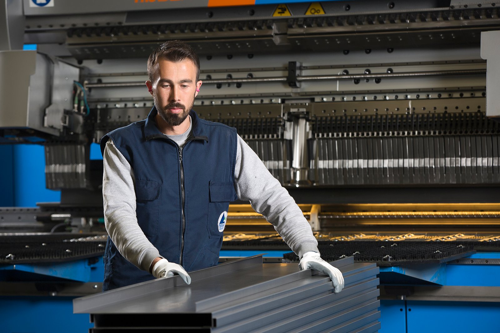Professional commercial industrial photographs by Sinan Niyazi Kutsal for Merih Elevator, capturing operators in navy blue company uniforms handling steel sheets at a bending machine and monitoring a laser cutting enclosure throwing bright sparks.