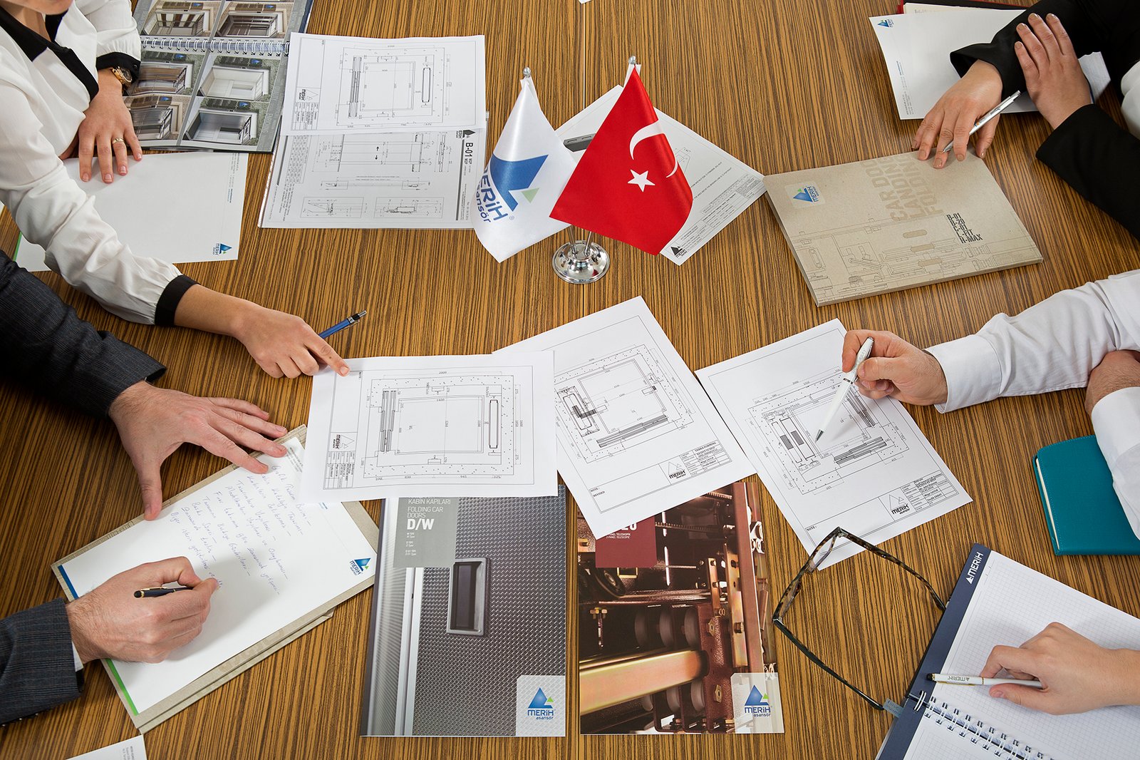 Professional commercial corporate photograph by Sinan Niyazi Kutsal for Merih Elevator, capturing a top-down view of hands pointing at elevator blueprints, technical folders, and catalogs around a Turkish desk flag during a boardroom meeting.