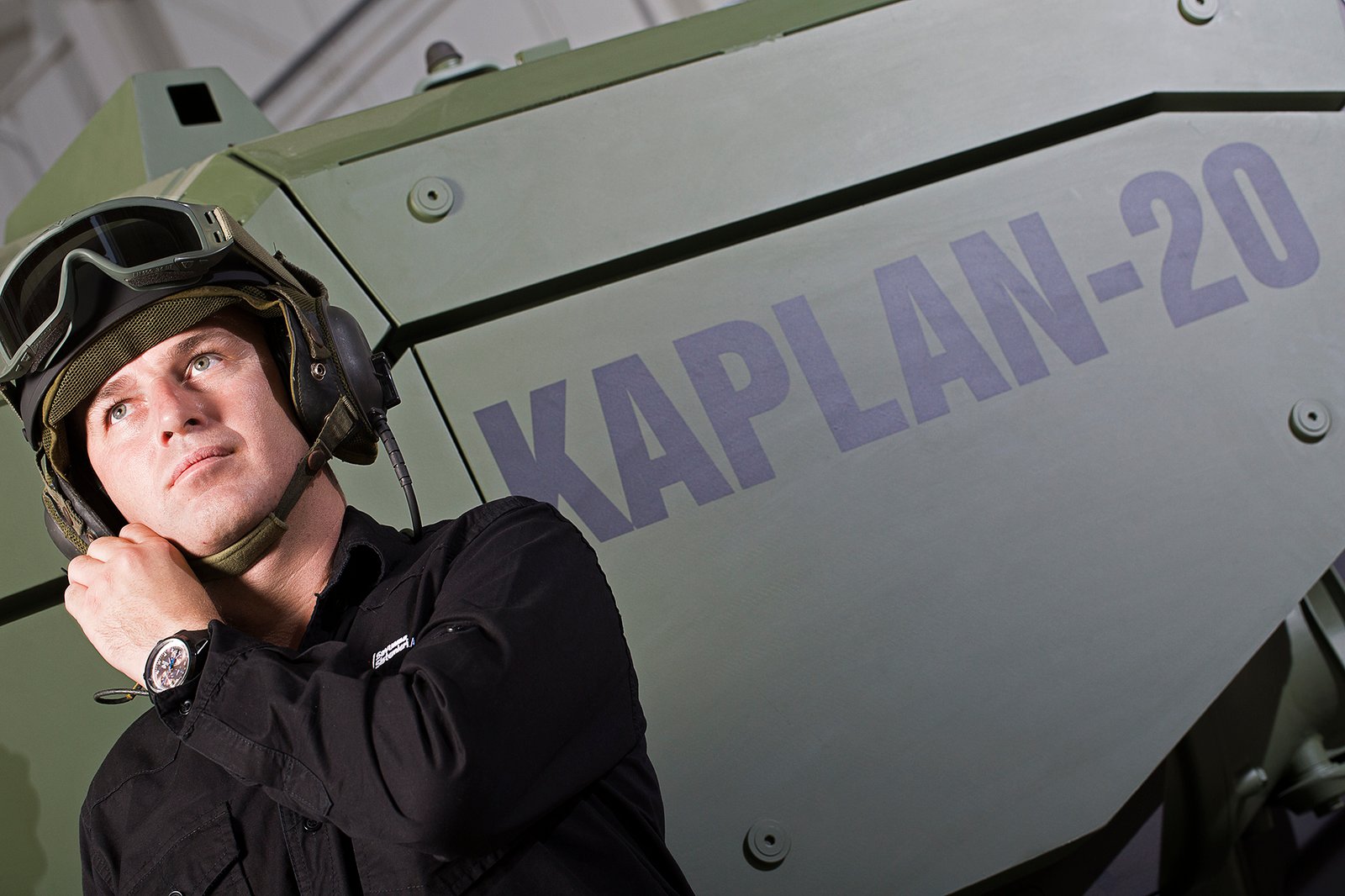 Professional commercial industrial photograph by Sinan Niyazi Kutsal for FNSS Defense Systems, capturing a military crew member in a tactical communication helmet and goggles posing in front of a green KAPLAN-20 armored combat vehicle.