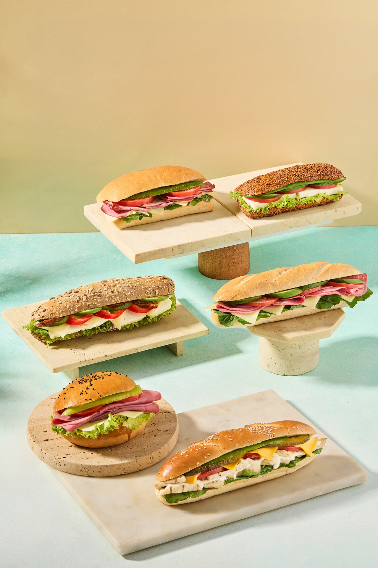 Studio photograph of assorted Simit Sarayı sandwiches arranged on display platforms with pastel backgrounds and hard daylight-style flash lighting.