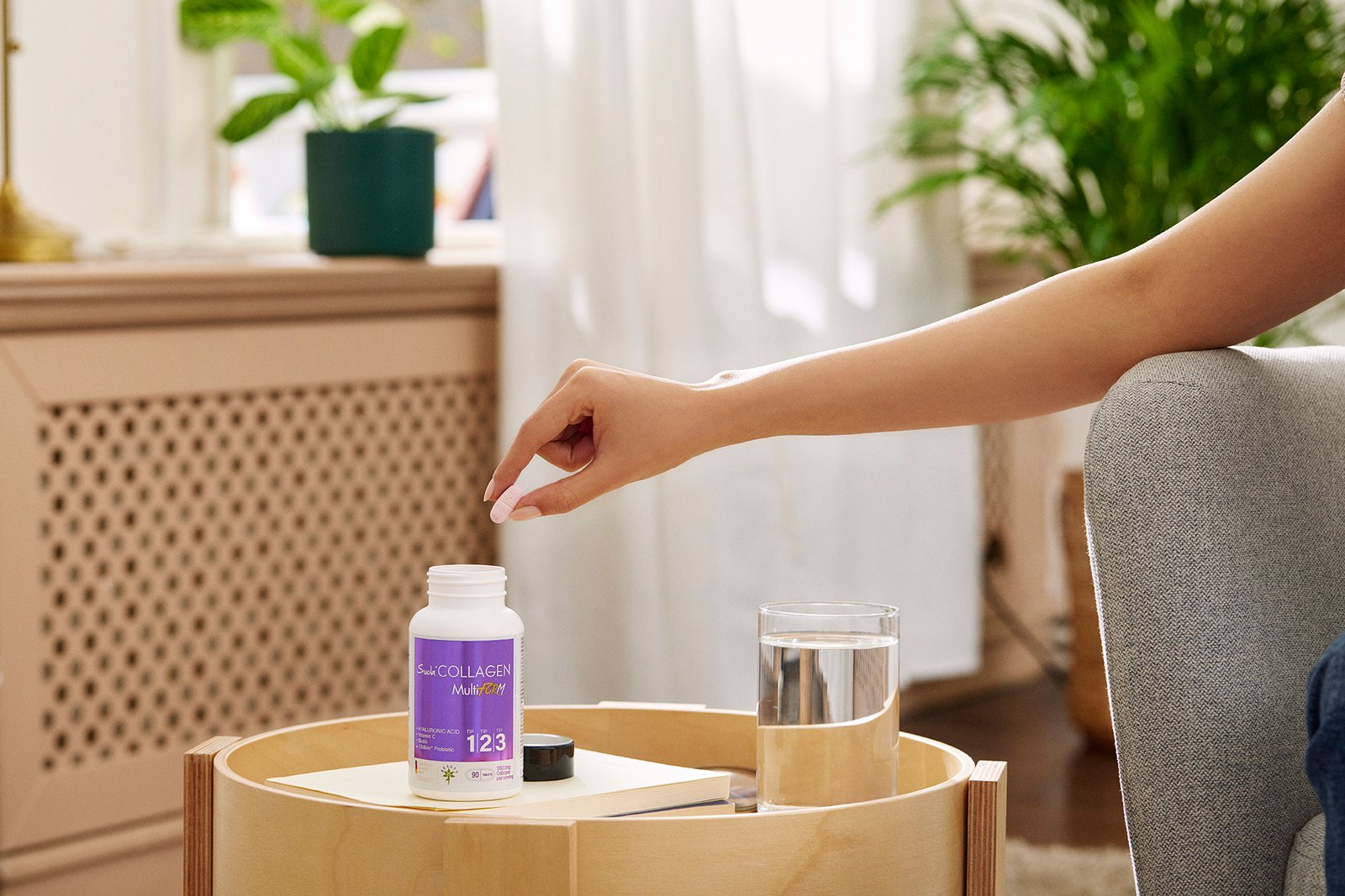 A professional commercial product photograph by Sinan Niyazi Kutsal capturing a close-up of a female hand taking a pink tablet from a Suda Collagen Multi Form bottle on a wooden table.
