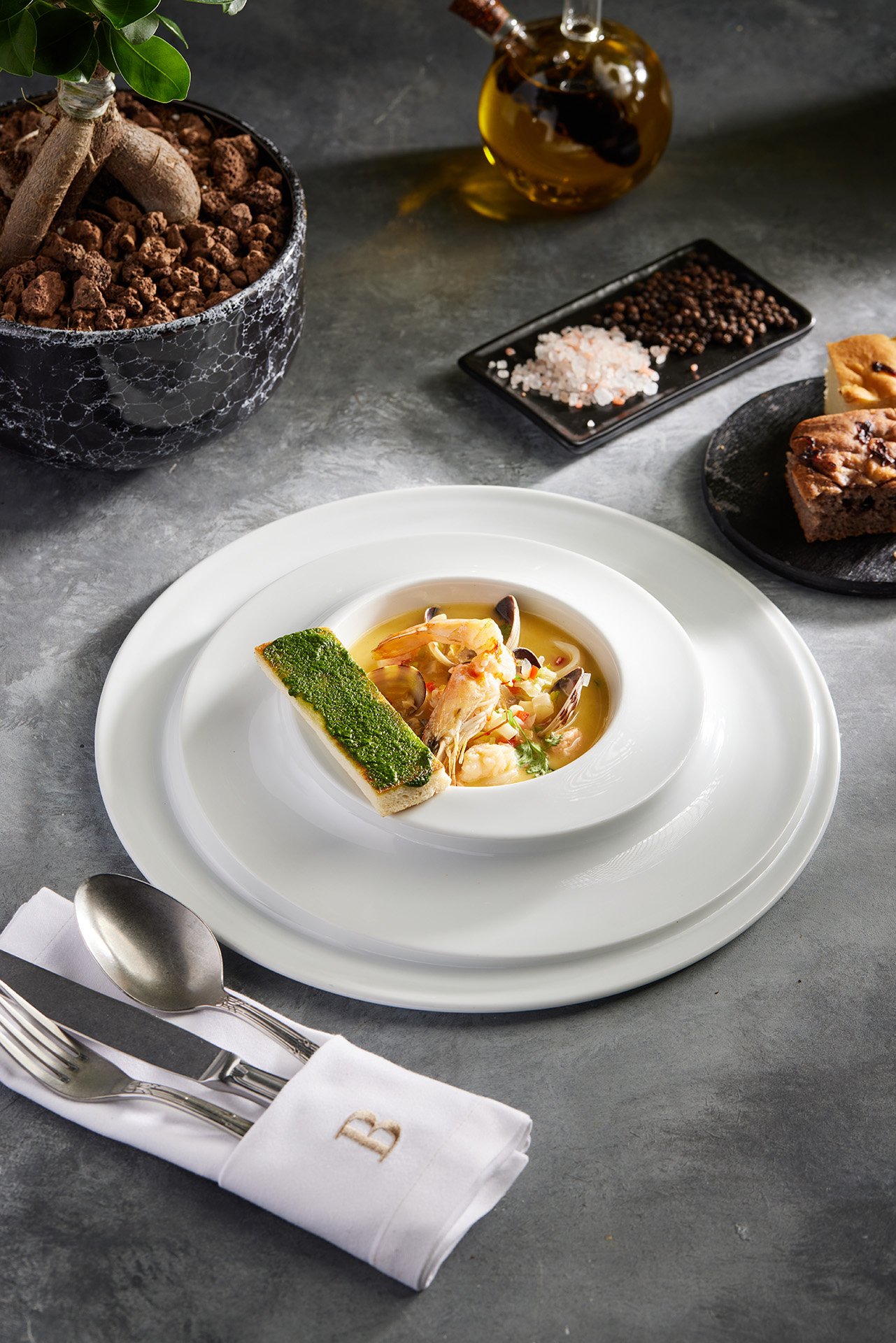 A beautiful commercial food photograph by Sinan Niyazi Kutsal for Barley Brasserie, capturing a luxury seafood soup with shrimp and clams served with a vibrant green pesto toast in a wide white bowl.