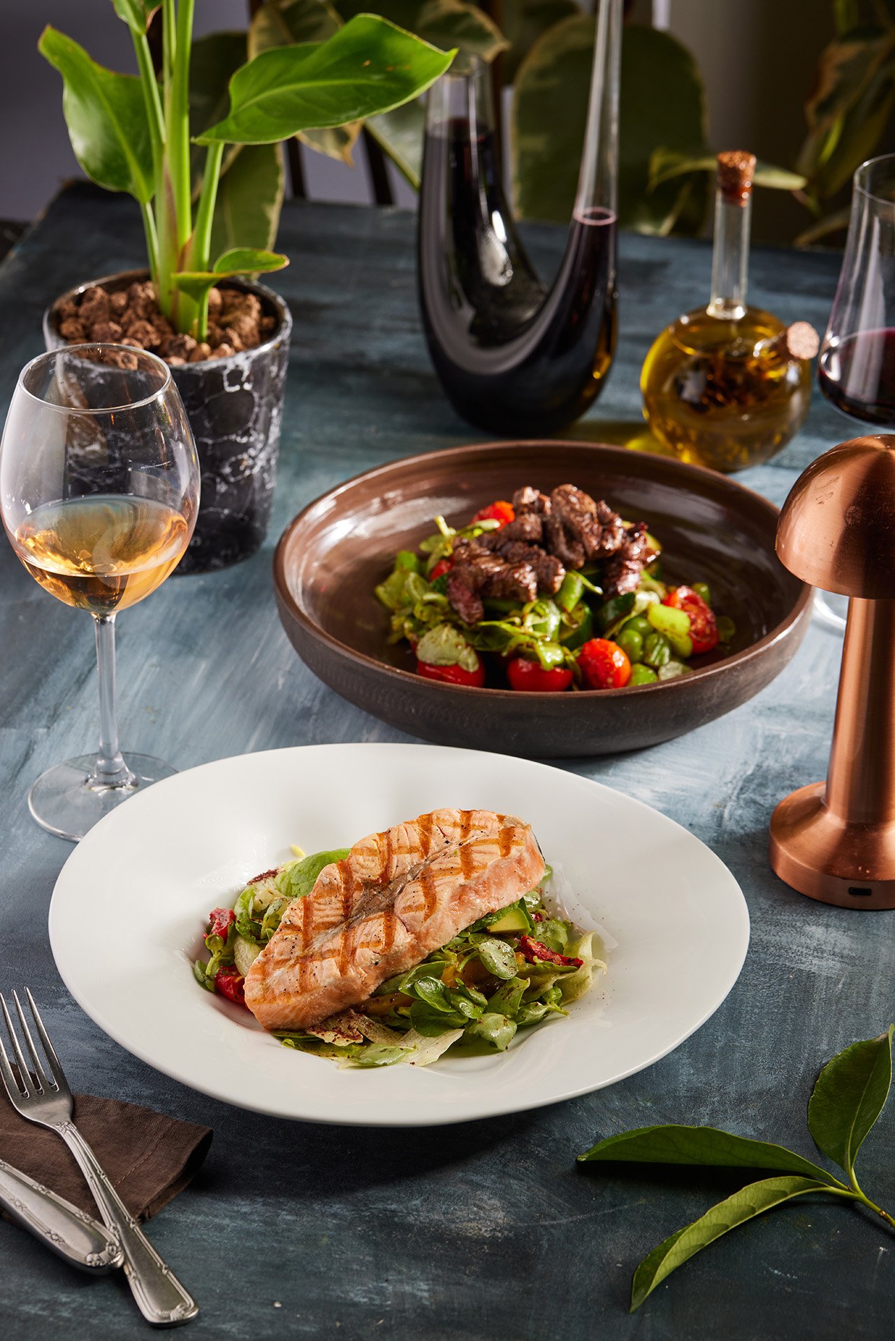A striking commercial food photograph by Sinan Niyazi Kutsal for Barley Brasserie, displaying a cross-hatched grilled salmon fillet in a white bowl and an upscale beef salad bowl on a styled dining table.
