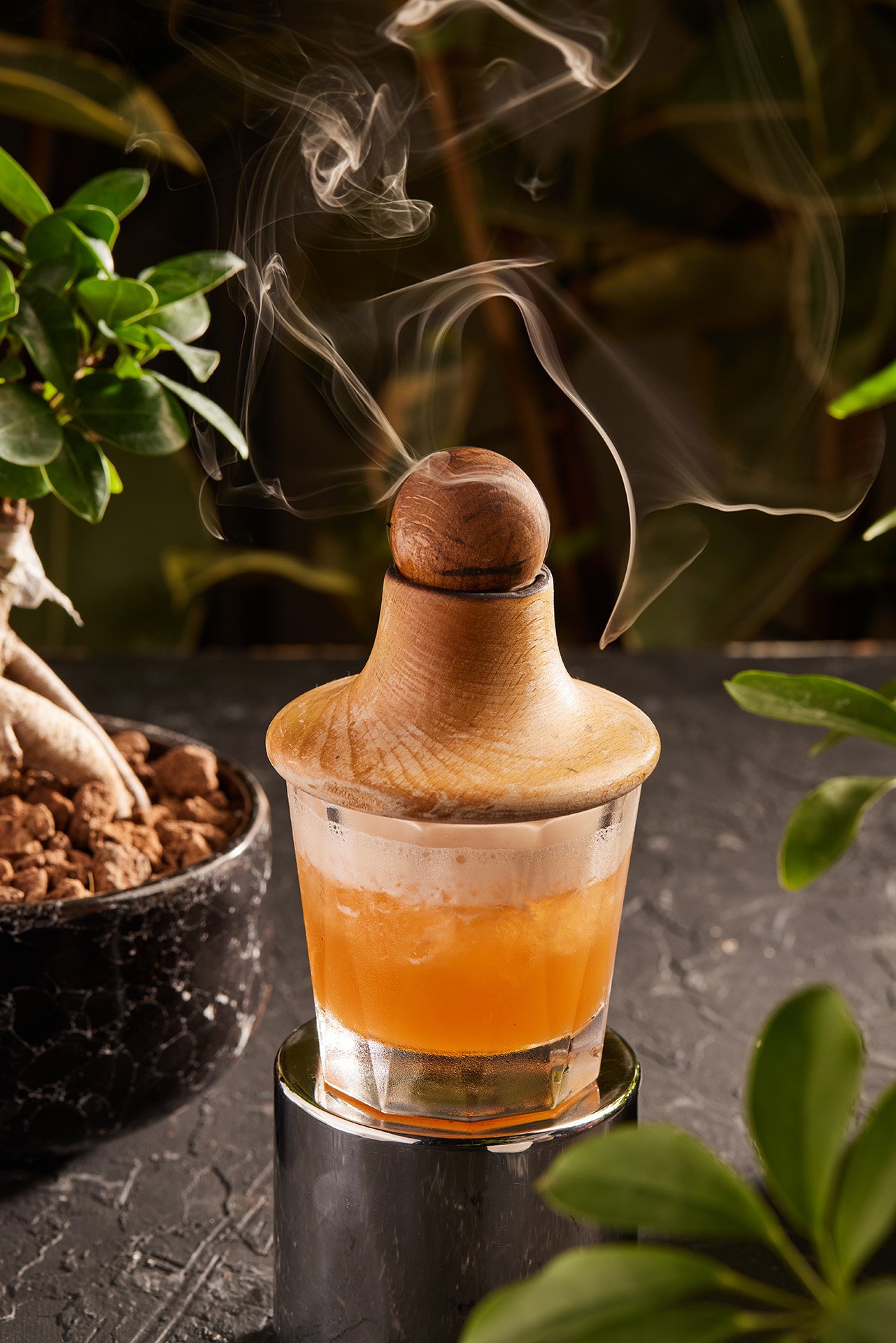 A captivating commercial beverage photograph by Sinan Niyazi Kutsal for Barley Brasserie, showcasing an amber cocktail inside a glass topped with a wooden smoking cap releasing beautiful wisps of smoke.