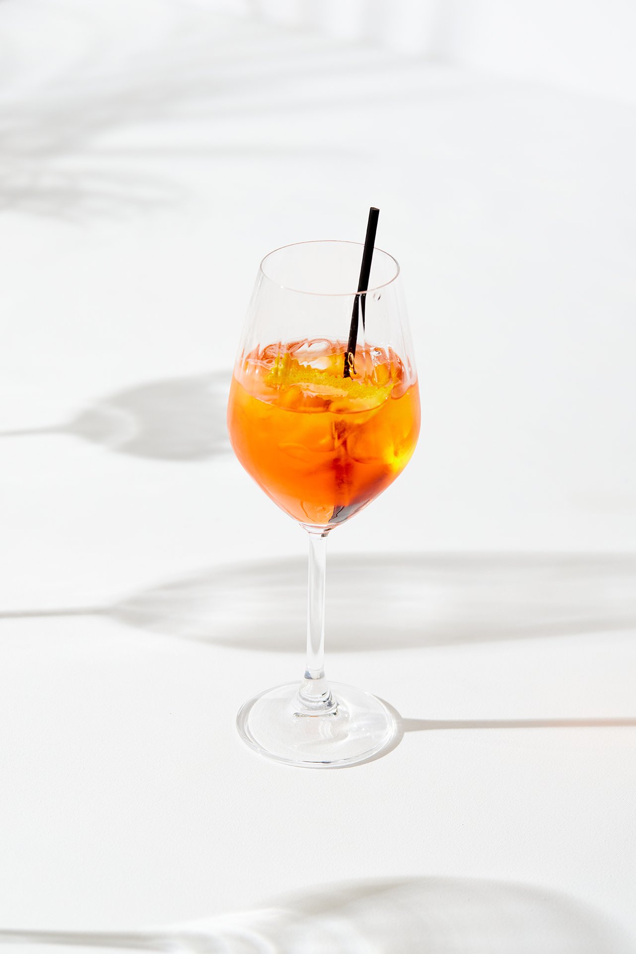 A flawless high-key commercial beverage photograph by Sinan Niyazi Kutsal for Barley Brasserie, featuring a bright orange Aperol Spritz cocktail in a stemmed wine glass with graphic shadows on a pure white background.