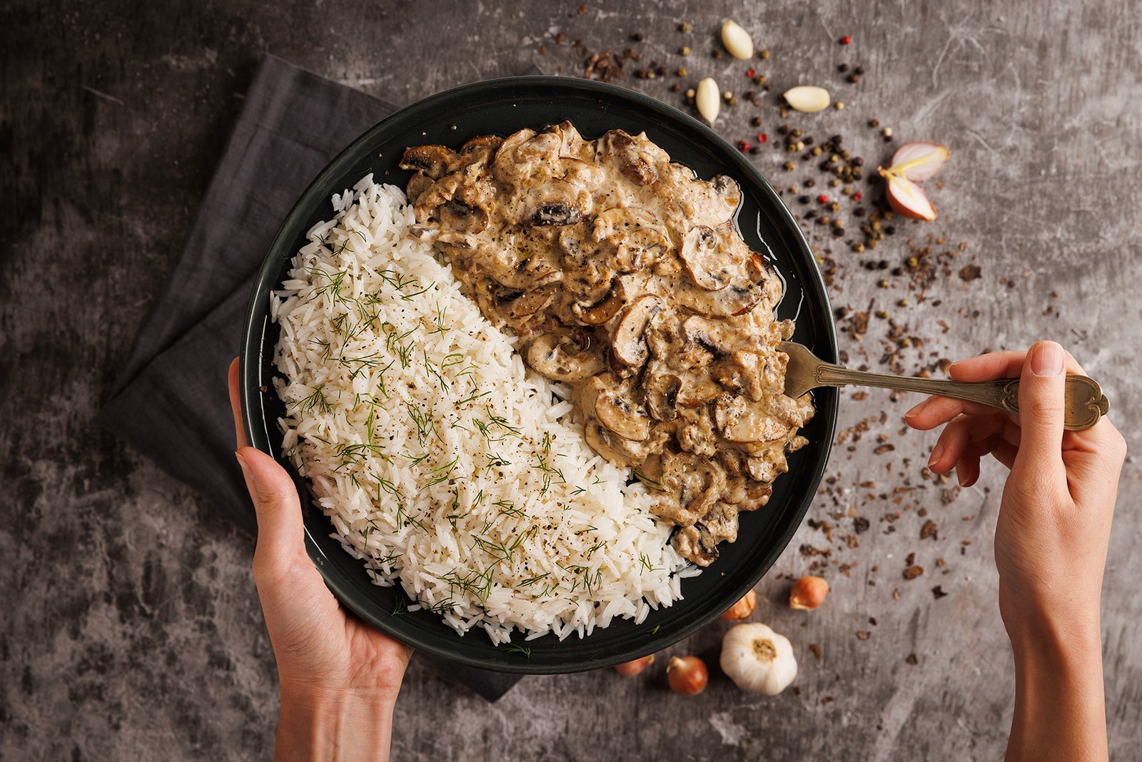 Professional commercial food advertising photograph by Sinan Niyazi Kutsal for Tarifte, capturing a top-down view of two hands holding a round black plate split between creamy mushroom beef stroganoff and white basmati rice with dill on a textured grey surface.