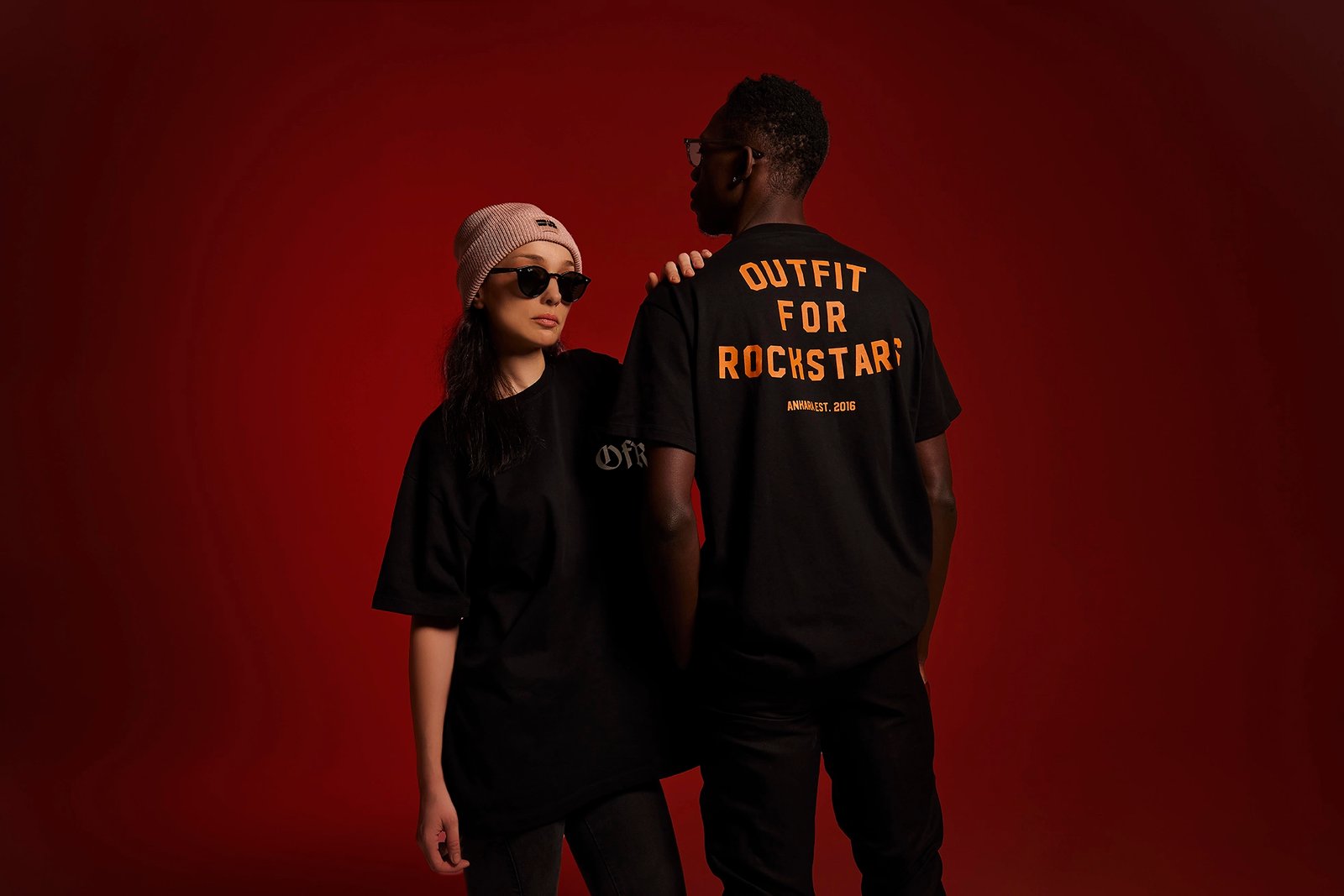 Fashion studio portrait by Sinan Niyazi Kutsal showcasing a black streetwear t-shirt with orange bold typography on the back reading 'OUTFIT FOR ROCKSTARS ANKRA EST. 2016', modeled by a male and female model against a dark red backdrop.