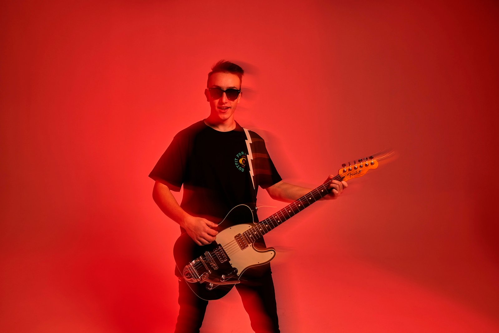 A commercial apparel photo by Sinan Niyazi Kutsal showing a young male model with sunglasses holding a black Fender electric guitar against a solid red studio background, captured with a subtle motion blur effect to emphasize a rock concert atmosphere.
