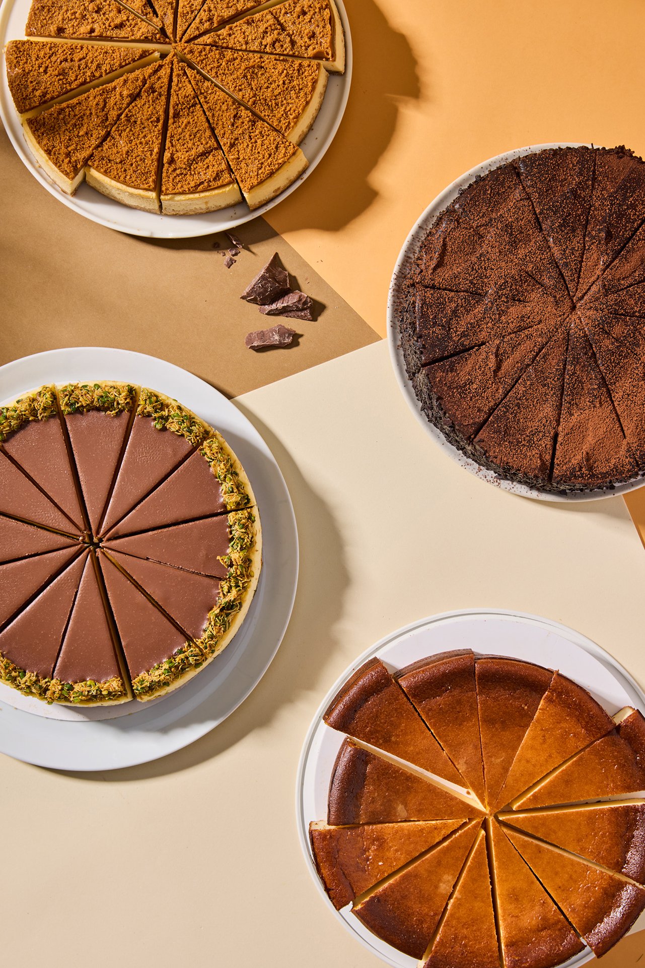Top-down studio photograph featuring a selection of Simit Sarayı cheesecakes arranged on plates with hard daylight-style flash lighting and geometric composition.