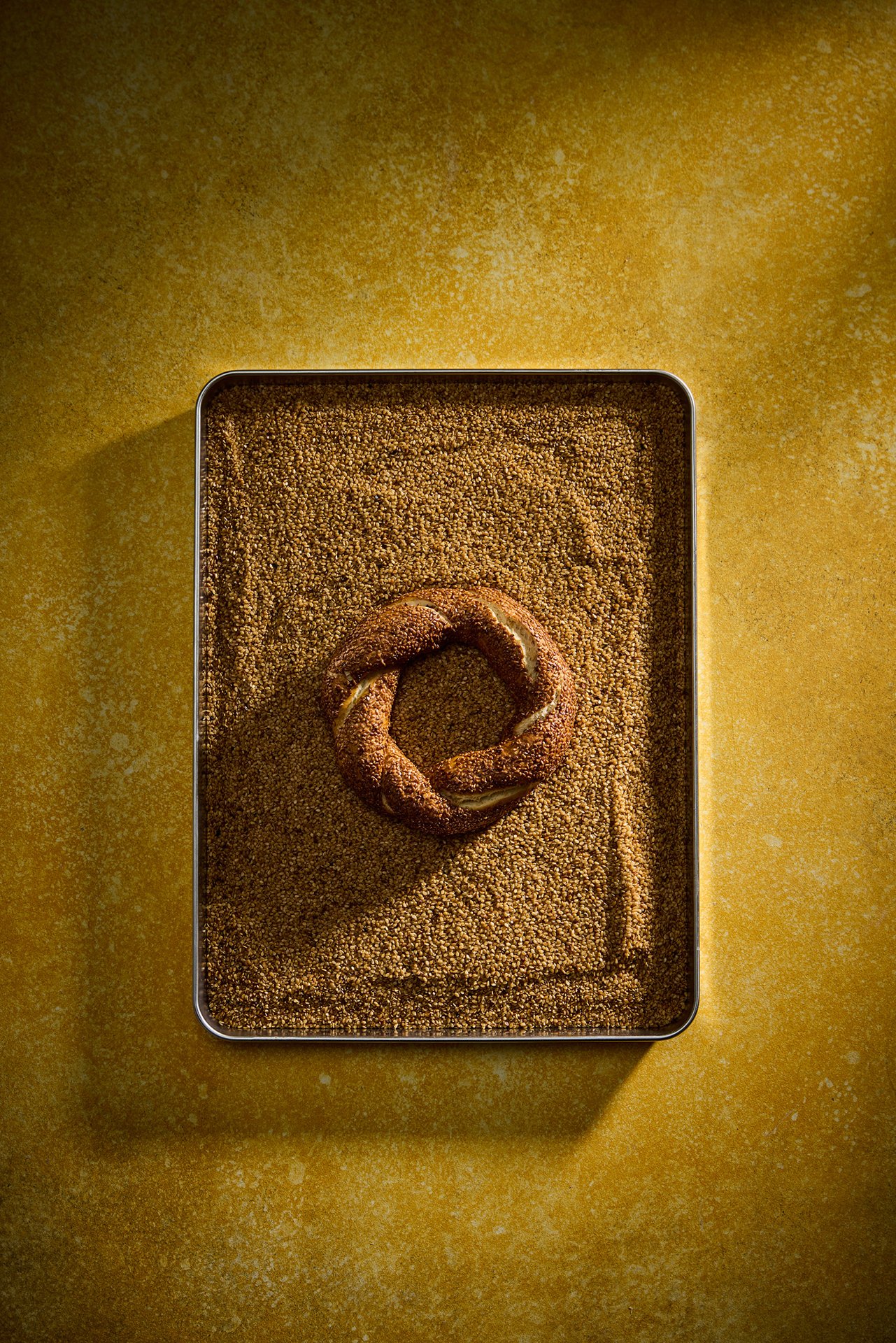 Top-down studio photograph of a single Simit Sarayı simit placed on a tray filled with sesame seeds, using hard daylight-style flash lighting.